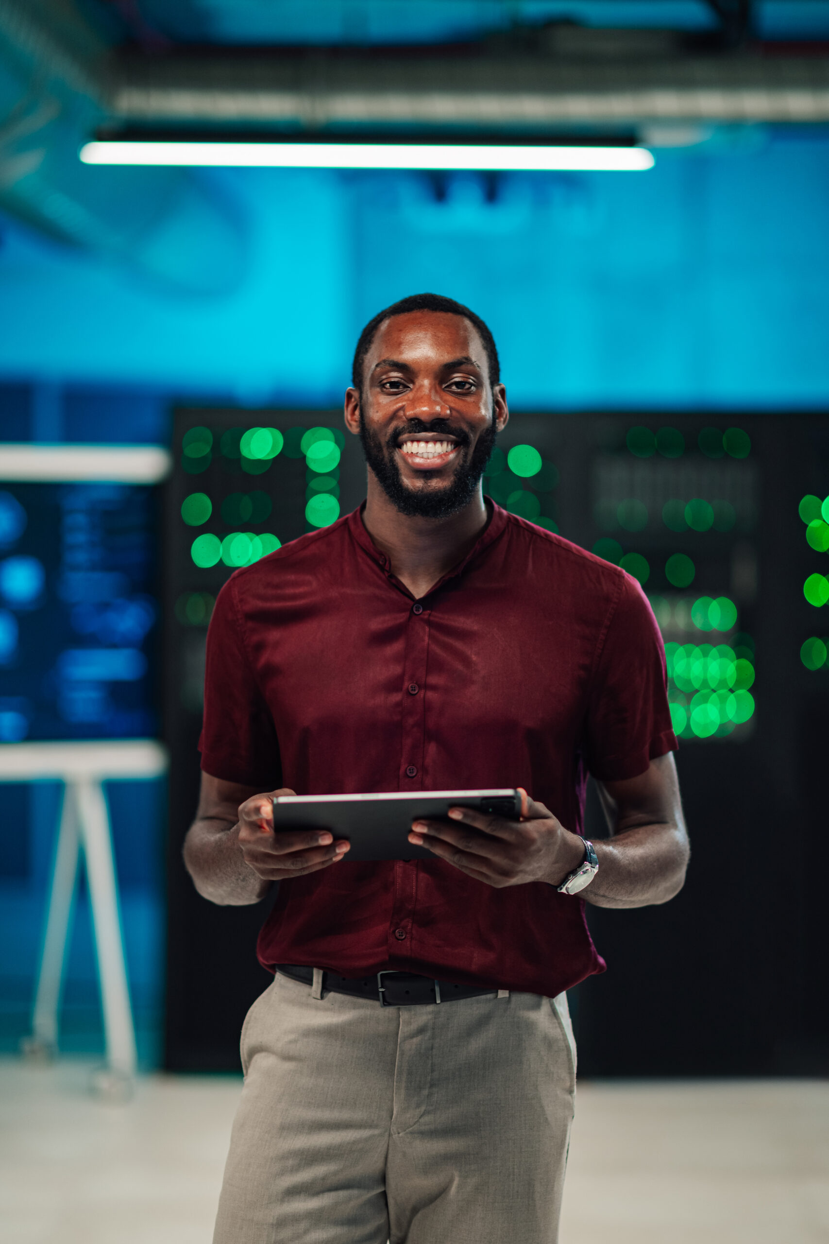 African american it technician smiling, holding a digital tablet in a server room, managing data and network